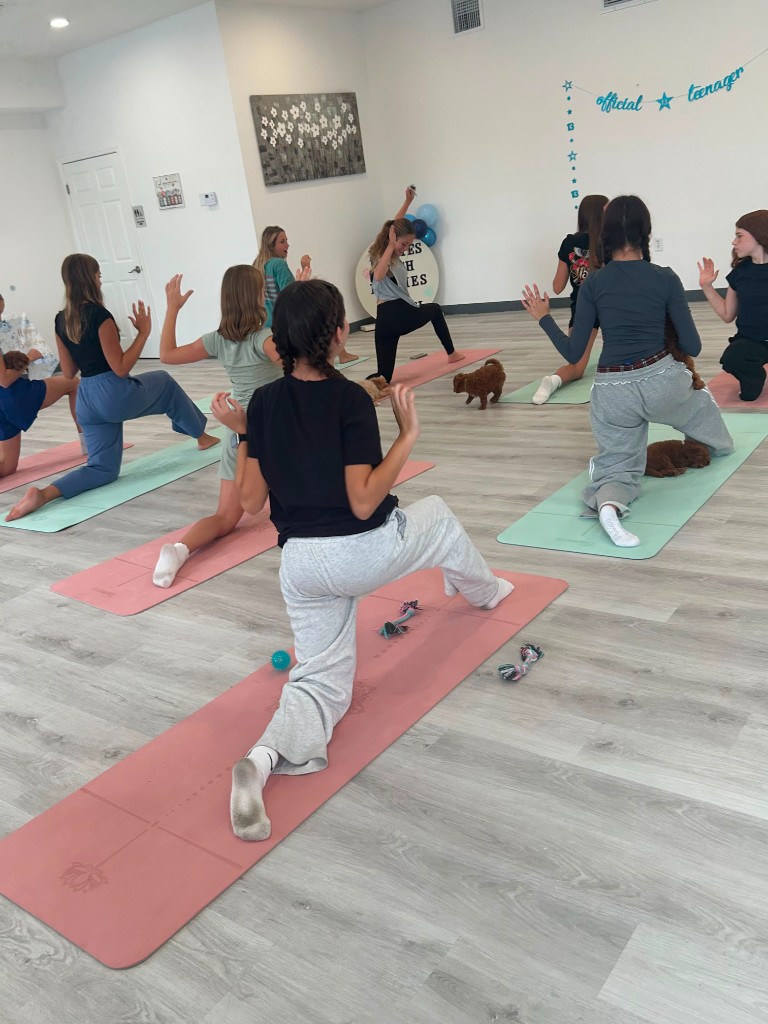 A cheerful 13th birthday Puppy Yoga session, with teen guests stretching on pink and mint mats while fluffy brown puppies roam the studio.