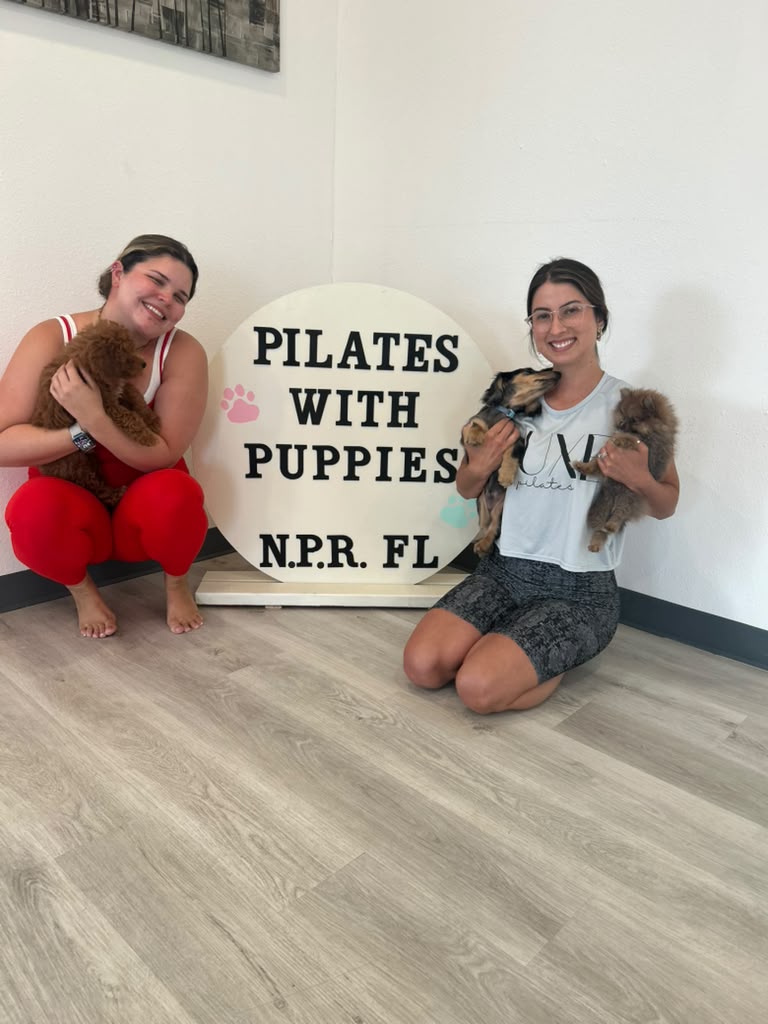 Two smiling guests posing with three puppies beside the circular Pilates with Puppies studio sign.