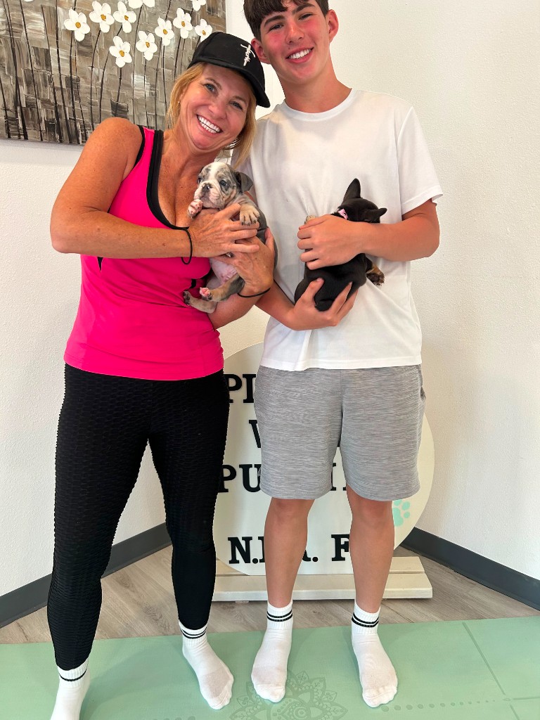 A woman and a teenage guest standing on a yoga mat, each holding a French Bulldog puppy in the studio.